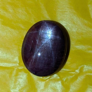 Puede incluir: Una gema pulida, de forma ovalada, de color burdeos intenso. La superficie presenta un brillo sutil y líneas internas visibles. La gema se encuentra sobre un fondo amarillo, resaltando su rico color y forma.