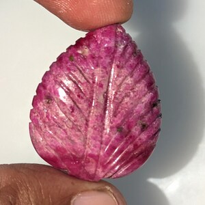 May include: A carved pink ruby gemstone in the shape of a leaf. The gemstone has a detailed leaf design and a smooth, polished surface.