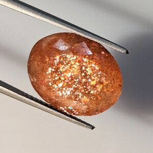 May include: A sunstone gemstone with a reddish-brown color and a faceted oval shape. The stone has a shimmery, iridescent quality.