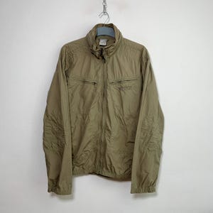 Può includere: Una giacca di colore marrone chiaro con chiusura a zip e due tasche sul davanti. La giacca ha un colletto alto e la parola "Nike" è ricamata sul lato destro del petto.