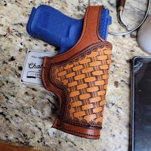 Puede incluir: Una funda de cuero marrón con un patrón de tejido de cesta y una pistola azul en su interior. La funda está cosida con hilo negro y tiene un borde decorativo.