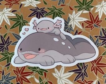 May include: A sticker featuring a cartoon axolotl character with a smaller axolotl on its back, both in shades of gray. The sticker has a white border and is set against a background of brown paper with colorful maple leaf designs.