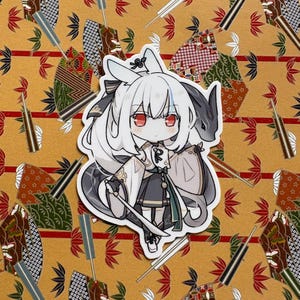 May include: A white sticker depicting an anime-style character with white hair, red eyes, and a black and grey outfit. The character is holding a sword. The background is a patterned yellow fabric with decorative elements.