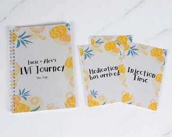 May include: A spiral-bound notebook and two cards with a white background and a pineapple design. The notebook reads "Lucie + Alex's IVF Journey". The cards say "Medication has arrived" and "Injection Time".