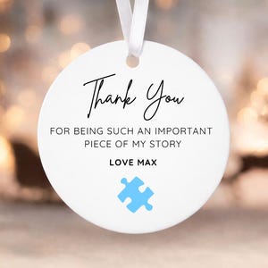 May include: A white, circular ornament with a hanging ribbon. It displays the words "Thank You" in cursive, a heartfelt message, and the name "Max." A blue jigsaw puzzle piece graphic is also present.