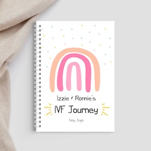 May include: A white spiral bound journal with a pink and orange rainbow design and the text "Izzie & Ronnie's IVF Journey" with a yellow starburst design. The text "Fortley Designs" is at the bottom of the journal.