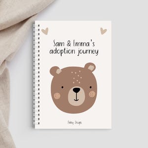 May include: A cream-coloured spiral-bound notebook with a cartoon bear face and the text "Sam & Emma's adoption journey." The bear is brown with blush-coloured cheeks and a black nose. Two small hearts are above the text.