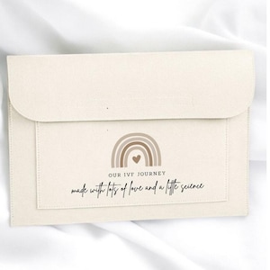May include: A cream-coloured felt document folder with a flap closure. The front features a printed graphic of a brown rainbow with a small heart in the centre, along with the text "OUR IVF JOURNEY" and "made with lots of love and a little science."