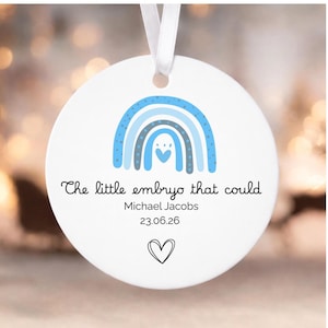 May include: A round white ceramic ornament with a white ribbon hanger. It features a blue rainbow graphic with a small heart, the text "The little embryo that could", the name "Michael Jacobs", the date "23.06.26", and a hand-drawn heart outline.