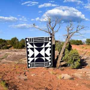 May include: A black and white geometric quilt with a star pattern is displayed against a backdrop of a desert landscape with a tree in the foreground.