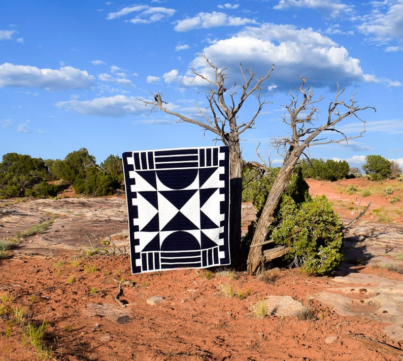 May include: A black and white geometric quilt with a star pattern is displayed against a backdrop of a desert landscape with a tree in the foreground.