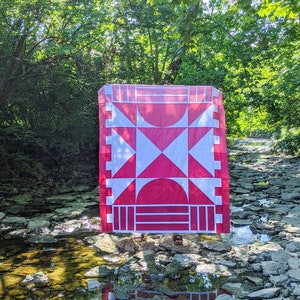 May include: A red and white quilt with a geometric pattern is displayed outdoors in a stream bed. The quilt is made of fabric squares and features a large red X in the centre.
