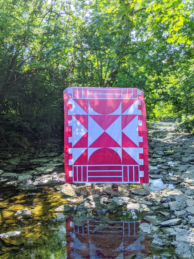 May include: A red and white quilt with a geometric pattern is displayed outdoors in a stream bed. The quilt is made of fabric squares and features a large red X in the centre.