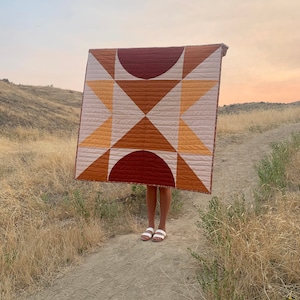 May include: A quilt with a geometric pattern in shades of orange, brown, and white. The quilt is being held up in front of a person standing on a dirt path.