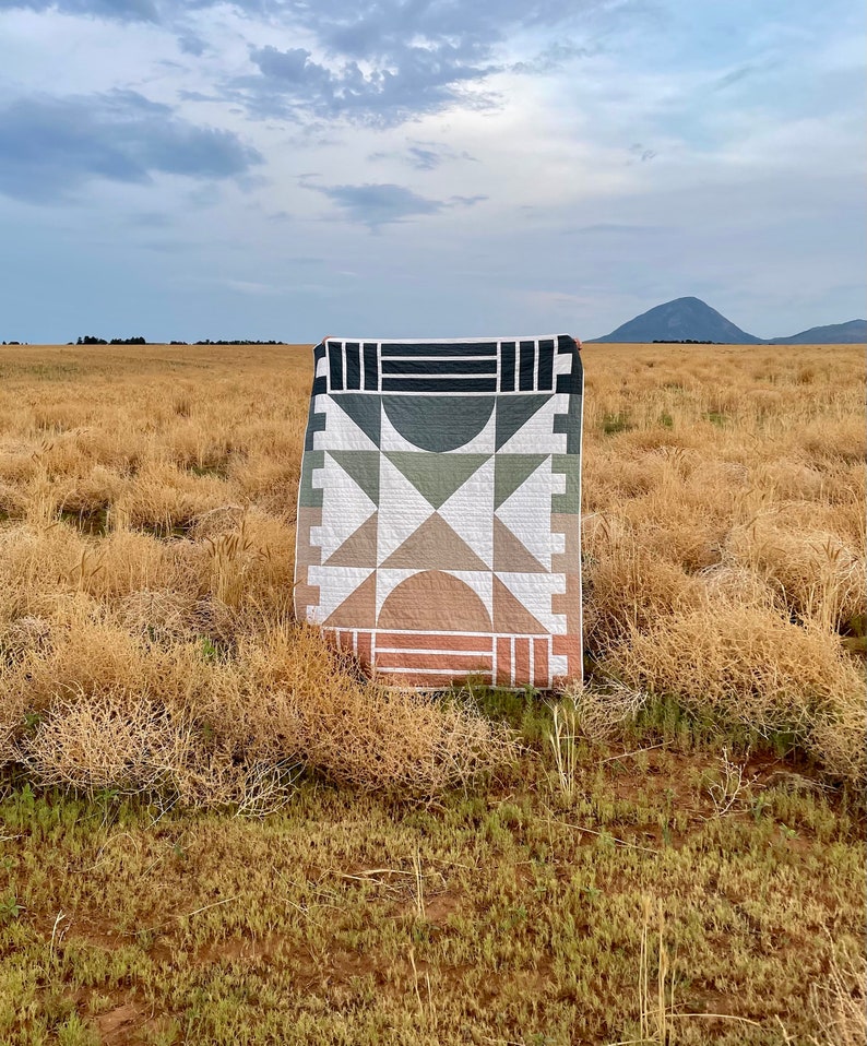 May include: A geometric quilt with a black, white, green, and brown colour scheme is displayed in a field of dry grass. The quilt is made up of squares and triangles.