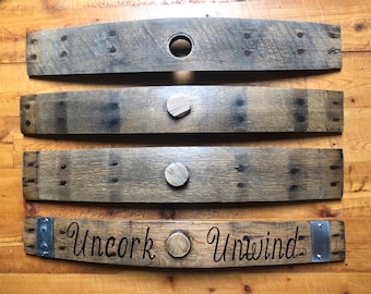 May include: Four reclaimed wood barrel staves. Three staves have wooden plugs. The bottom stave has the words "Uncork Unwind" in a script font, with metal brackets on each end. The wood is a weathered gray color.