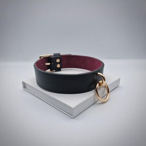 Puede incluir: Collar de cuero negro con interior burdeos y herrajes dorados. El collar presenta un cierre de hebilla y un gran anillo dorado. El collar está sobre una superficie blanca.