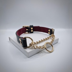 May include: Black leather collar with a burgundy interior, gold-colored hardware, and a gold chain detail. The collar features a buckle closure and two gold rings. The collar is sitting on a white surface.