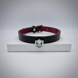 May include: Black leather collar with a burgundy interior. The collar features a silver-colored D-ring and buckle closure. The collar is displayed on a white surface.