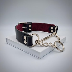 May include: Black leather collar with a burgundy interior, featuring silver-tone hardware. The collar has a buckle closure and is adorned with a chain and two rings. The item is displayed on a white surface.