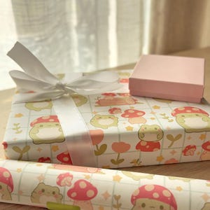 May include: Two gift boxes wrapped in white paper with a frog and mushroom pattern. The larger box has a white satin ribbon tied in a bow. The smaller box is pink and is sitting on top of the larger box.