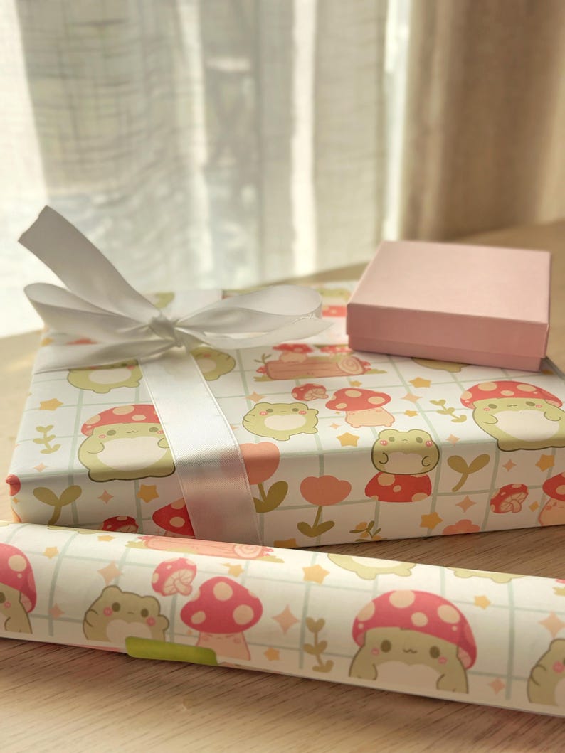 May include: Two gift boxes wrapped in white paper with a frog and mushroom pattern. The larger box has a white satin ribbon tied in a bow. The smaller box is pink and is sitting on top of the larger box.