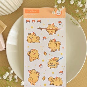 May include: A sticker sheet with cartoon characters in different poses, set against a light orange backdrop. The stickers feature the text "POMBON" and "STICKER SHEETS". The sheet is placed on a white plate, with a floral arrangement.