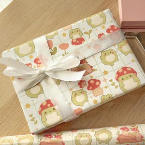 May include: A white gift box with a white ribbon wrapped around it. The box is covered in a pattern of green frogs, red mushrooms, and yellow stars on a white background.