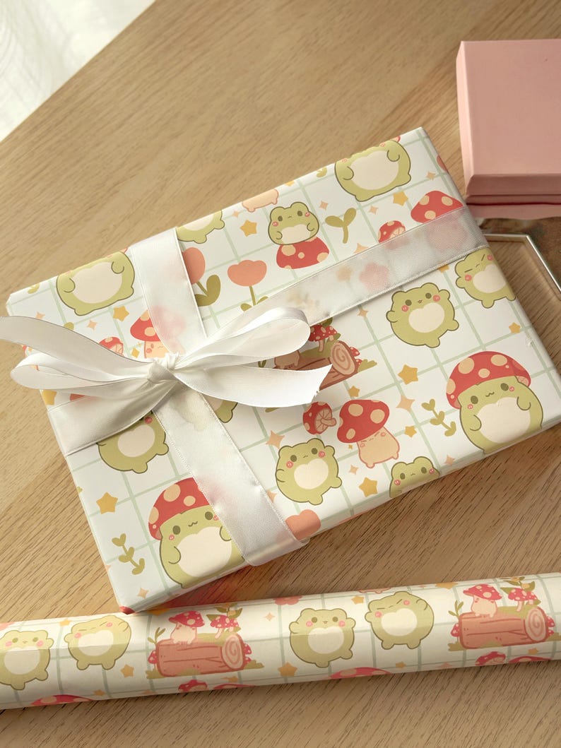 May include: A white gift box with a white ribbon wrapped around it. The box is covered in a pattern of green frogs, red mushrooms, and yellow stars on a white background.