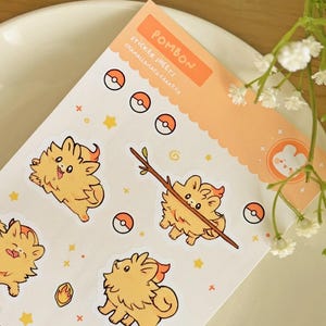 May include: A sticker sheet with cartoon character stickers. The stickers feature an orange, fluffy character with a flame-like mane, stars, and Pok&eacute; Ball designs. The sheet has the word "POMBON" and "STICKER SHEETS" on an orange background.