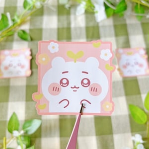 Puede incluir: Una pegatina cuadrada rosa con un personaje blanco con forma de oso, mejillas sonrojadas, ojos grandes y un brote. La pegatina está decorada con flores y corazones amarillos. Dos pegatinas adicionales son visibles en el fondo.