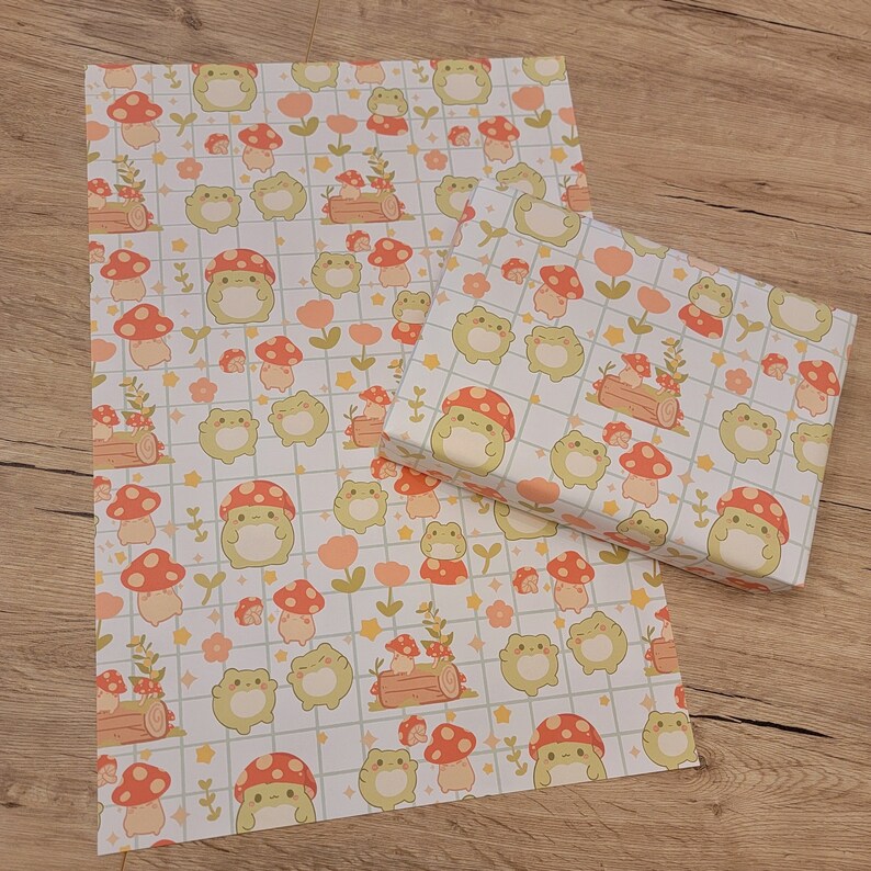 May include: Two sheets of wrapping paper with a repeating pattern of green frogs, orange mushrooms, and white flowers on a white and light brown grid background.