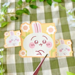 Puede incluir: Tres pegatinas con un conejito de dibujos animados con una expresión triste. El conejito es blanco con detalles en rosa, rodeado de flores amarillas y blancas. Las pegatinas están sobre un fondo a cuadros verde y blanco.