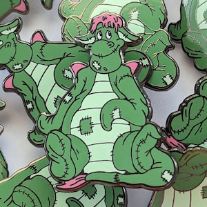 May include: A set of green and pink enamel pins featuring a cartoon dragon with stitched seams. The dragon has a pink mohawk and a friendly expression.