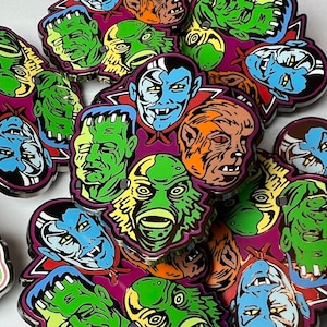Puede incluir: Una colección de pines de esmalte coloridos con varios personajes de monstruos, incluyendo Frankenstein, Drácula, la criatura de la laguna negra y un hombre lobo. Los pines son de diferentes tamaños y formas, y están dispuestos en un patrón aleatorio.