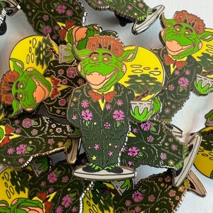 May include: Enamel pins featuring a green creature with orange hair, a red bow tie, and a floral suit, holding a small potted plant. The pins have a yellow moon background and are scattered across the image.