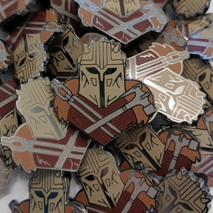 May include: A close-up of a pile of enamel pins featuring a cartoon character wearing a helmet and holding a weapon. The pins are in shades of brown, red, and silver.
