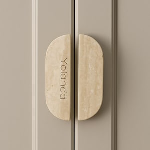 May include: Two cabinet door handles with a light beige, textured finish. Each handle is a half-oval shape with rounded ends. The word "Yolanda" is engraved vertically on one handle. The cabinet doors are a neutral grey colour.