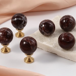 Può includere: Sei pomelli decorativi per mobili. I pomelli hanno un design sferico in marmo rosso scuro con venature bianche, montati su una base color oro. Alcuni pomelli sono appoggiati su una pietra quadrata bianca. Lo sfondo include un tessuto color pesca.