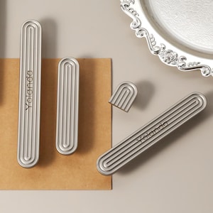 May include: A collection of silver-toned cabinet pulls and knobs with a grooved design. The word "Yolanda" is engraved on the pulls. A decorative silver tray and a green notebook are also in the image. The cabinet hardware is on a neutral background.