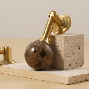 May include: A decorative cabinet knob with a gold-colored base and a brown marble sphere. The knob is displayed on a light-colored stone block, showcasing its design and texture. The background is a neutral beige.