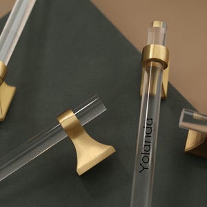 May include: Clear acrylic cabinet pulls with gold-toned metal accents. The pulls have a cylindrical design with a gold band at each end, and a gold-toned base. The word "Yolanda" is printed on one of the pulls.