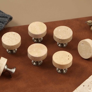 Op de afbeelding: Een set van zeven ronde kastknoppen met een textuurrijk, beige stenen bovenoppervlak gemonteerd op een metalen zilverkleurige basis. Elke knop heeft een plat, rond voorvlak met natuurlijke, poreuze details voor een consistent uiterlijk.