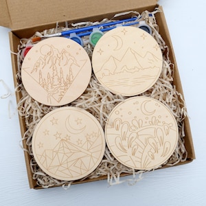 Op de afbeelding: Een set van vier onafgewerkte houten cirkels met berg- en maanmotieven, klaar om te worden geschilderd. De set bevat verf en kwasten.