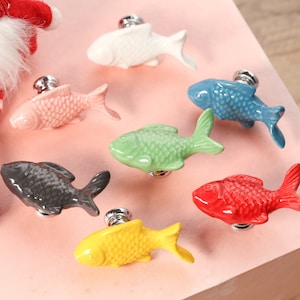 Può includere: Set di sette tiretti per cassetti in ceramica a forma di pesce in vari colori, tra cui bianco, rosa, blu, verde, grigio, rosso e giallo. Ogni tiretto presenta un design di pesce realistico con un pomello in metallo argentato.