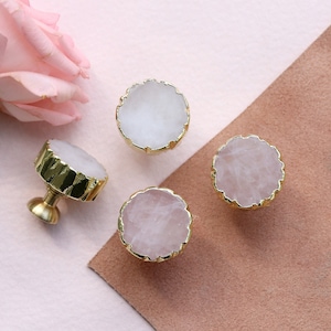 Pode incluir: Quatro puxadores de gaveta dourados com uma pedra redonda, translúcida, branca e semelhante a um cristal no centro. A pedra tem uma borda áspera e irregular. Os puxadores estão sobre uma superfície rosa e marrom.