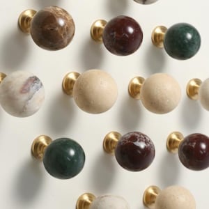 Può includere: Una collezione di pomelli sferici per mobili con varie finiture in pietra naturale, tra cui marmo marrone, rosso scuro, verde e crema, ciascuno montato su una base in ottone lucido, disposti a griglia su uno sfondo bianco uniforme.