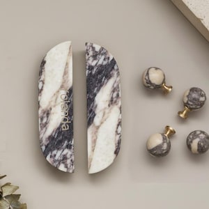 May include: A selection of marble cabinet hardware, featuring two large, half-moon shaped pulls and four small, spherical knobs. The pulls and knobs display a mix of white, grey, and black veining. The pulls have the word "olenda" printed on them.