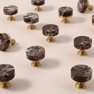 May include: A collection of circular cabinet knobs featuring a dark brown marble top with white veining and a polished brass base. Each knob has a flat, round face and a short, cylindrical stem that connects to a circular mounting plate.