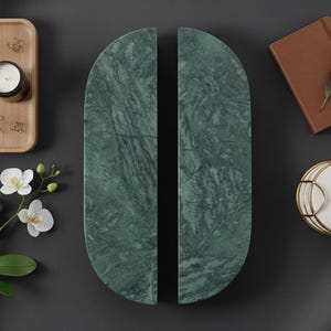 May include: A flat lay featuring decorative objects on a dark grey backdrop. It includes a wooden tray with a candle and paperclips, two green marble panels, a small brown book with a feather, and white marble coasters with gold edging.